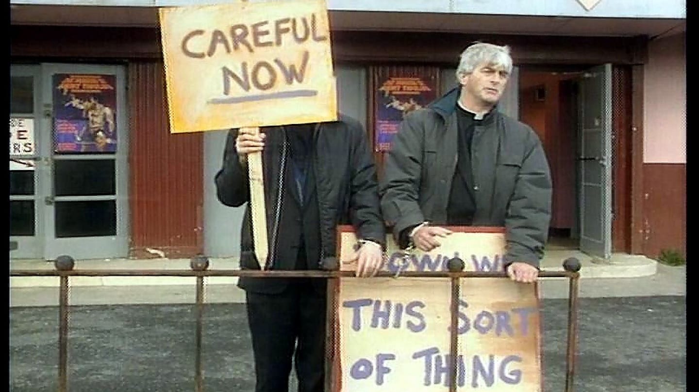 Father Crilly and Father McGuire handcuffed to a fence with protest placards from TV show 'Father Ted'