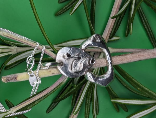 Silver jewellery pendant resembling Edvard Munch's 'The Scream' top of a sprig of rosemary green background.